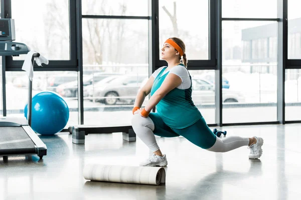 Plus size woman in sportswear stretching near fitness mat and treadmill — Stock Photo