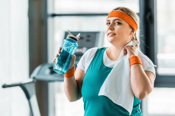 Exhausted plus size girl holding bottle with water and towel in gym — Stock Photo