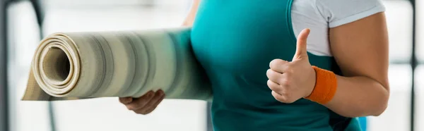 Vista recortada de la mujer con sobrepeso mostrando el pulgar hacia arriba mientras sostiene la estera de fitness - foto de stock