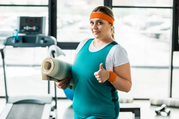 Glückliche übergewichtige Frau zeigt Daumen nach oben, während sie Fitnessmatte im Fitnessstudio hält — Stockfoto