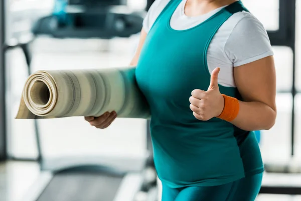 Ausgeschnittene Ansicht einer übergewichtigen Frau, die den Daumen nach oben zeigt, während sie eine Fitnessmatte in der Turnhalle hält — Stockfoto