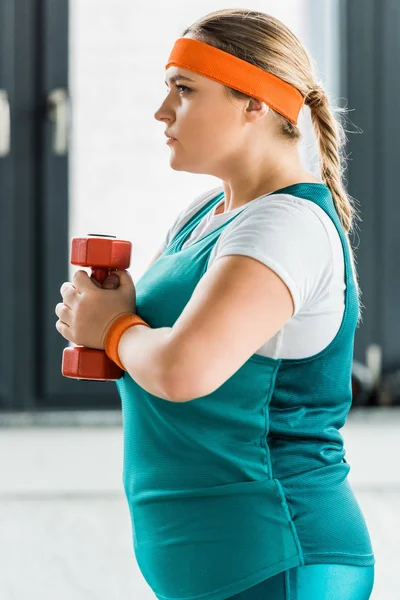 Vista lateral de la chica con sobrepeso grave sosteniendo la mancuerna en el gimnasio - foto de stock