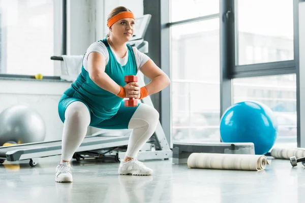 Happy overweight girl squatting with dumbbell in gym — Stock Photo