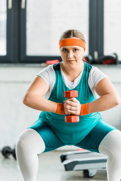 Attraente ragazza in sovrappeso in abbigliamento sportivo accovacciato con manubri in palestra — Foto stock