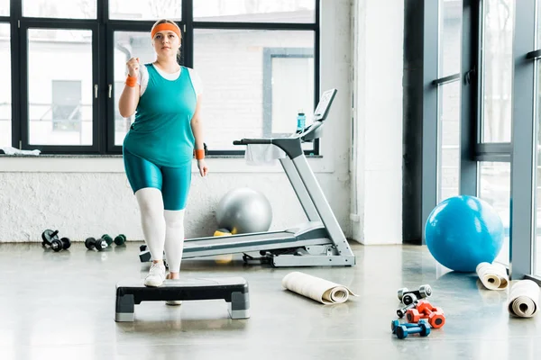 Attractive plus size girl standing on step platform near dumbbells in gym — Stock Photo