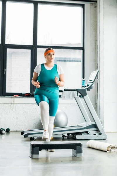 Plus size woman exercising on step platform near fitness mat in gym — Stock Photo