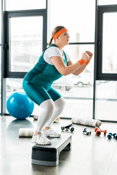 Plus size woman squatting on step platform near fitness mats and dumbbells — Stock Photo