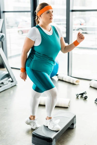 Más tamaño chica ejercitando en paso plataforma cerca de colchonetas de fitness en el gimnasio - foto de stock