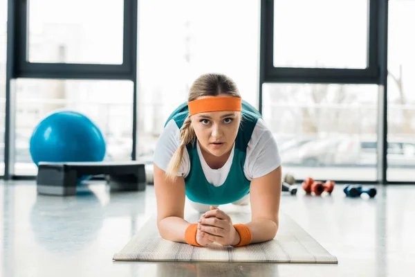 Menina com sobrepeso fazendo exercício prancha no tapete de fitness e olhando para a câmera — Fotografia de Stock