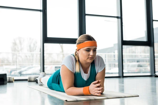 Pensive overweight girl relaxing on fitness mat in gym — Stock Photo
