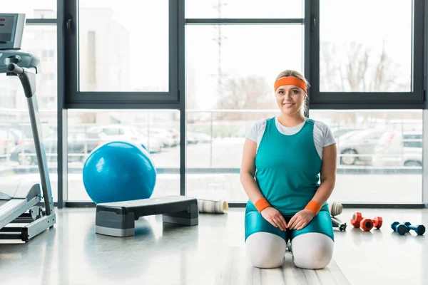 Alegre mulher plus size sentado no tapete de fitness perto de equipamentos desportivos — Fotografia de Stock