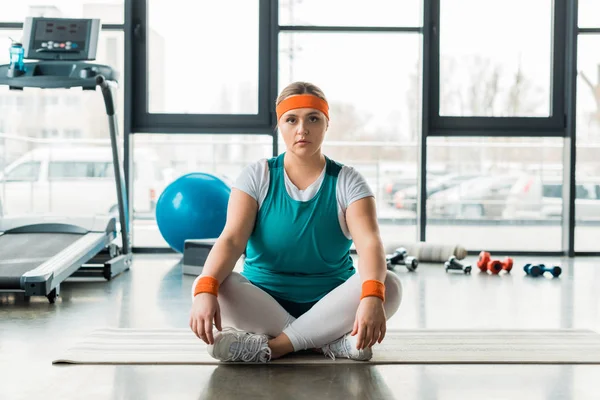 Сфокусированный плюс размер девушки, сидящей на фитнес-мат возле спортивного оборудования со скрещенными ногами — стоковое фото