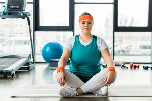 Más tamaño mujer sentada en la colchoneta de fitness cerca de equipo deportivo con las piernas cruzadas y los ojos cerrados - foto de stock