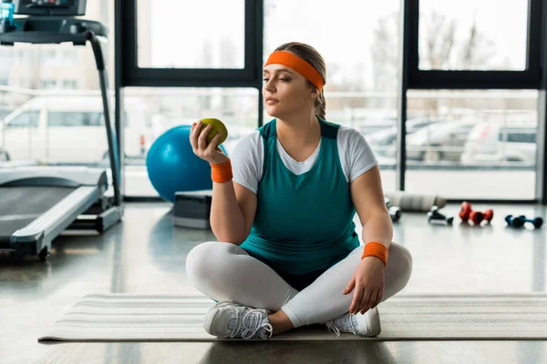 Plus size woman sitting on fitness mat near sport equipment with crossed legs and looking at apple — Stock Photo