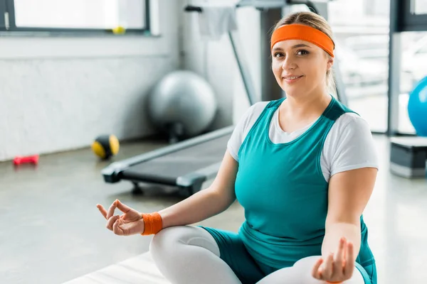 Selective focus of happy plus size woman sitting on fitness mat with crossed legs — Stock Photo