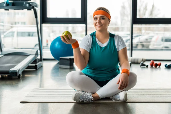 Плюс размер женщина сидит на фитнес-мат возле спортивного оборудования со скрещенными ногами и держа зеленое яблоко — стоковое фото
