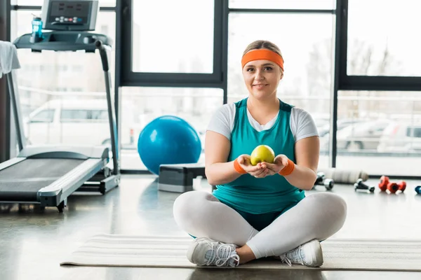 Além de mulher tamanho sentado no tapete de fitness perto de equipamentos desportivos com pernas cruzadas e segurando maçã — Fotografia de Stock