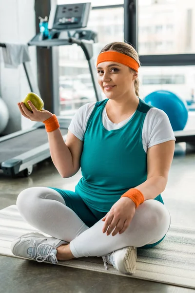 Happy plus size girl sitting on fitness mat with crossed legs and holding tasty apple — Stock Photo