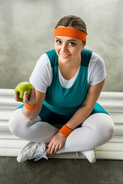Над головой вид счастливой плюс размер девушка сидит на фитнес-коврик со скрещенными ногами и проведение вкусное яблоко — стоковое фото