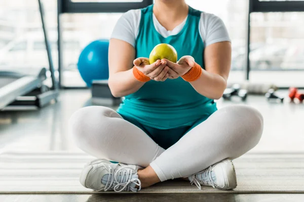 Cropped view of plus size girl sitting on fitness mat with crossed legs and holding tasty apple — Stock Photo