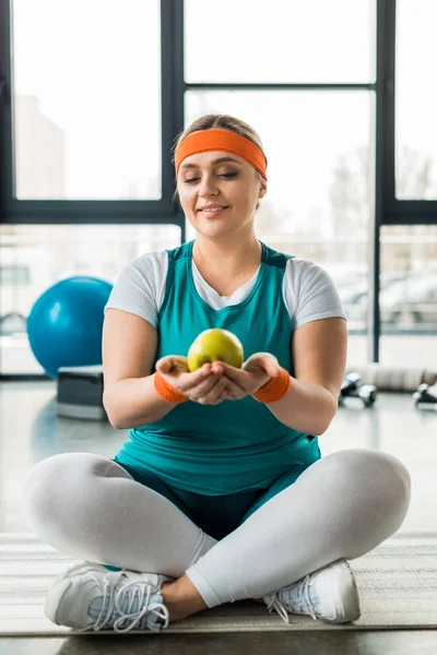 Happy plus size woman sitting on fitness mat with crossed legs and looking at tasty apple — Stock Photo