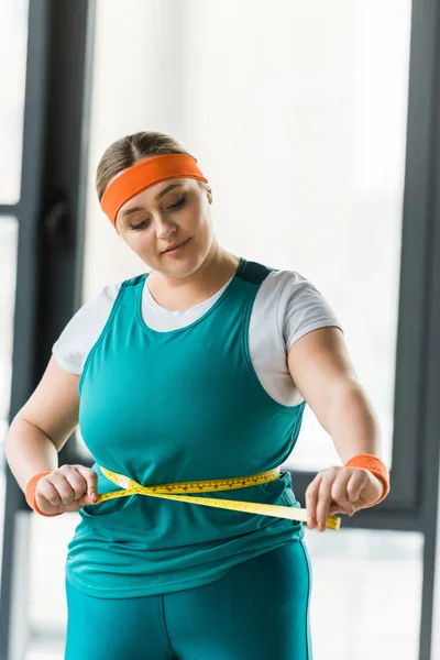 Plus Size Girl schaut auf Maßband beim Messen der Taille im Fitnessstudio — Stockfoto