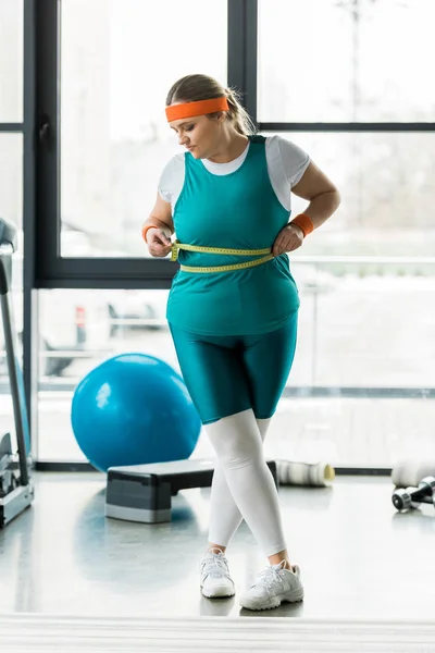 Menina com sobrepeso olhando para fita métrica ao medir a cintura no ginásio — Fotografia de Stock