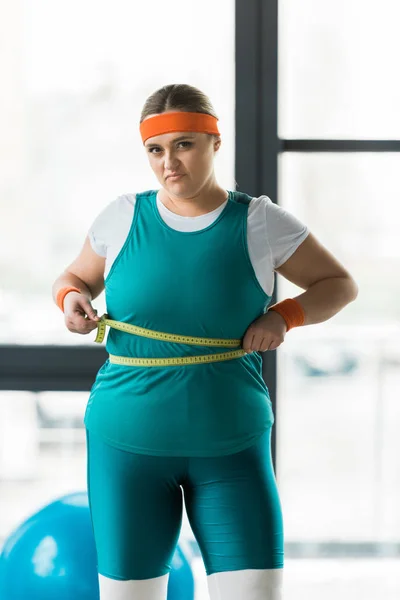 Chica con sobrepeso insatisfecha que mide la cintura en el gimnasio - foto de stock