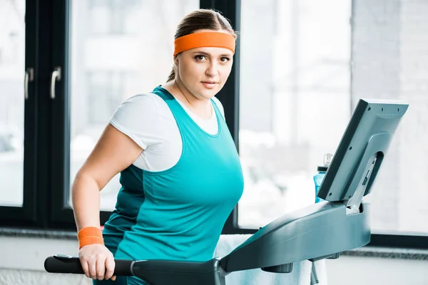 Menina com sobrepeso atraente se exercitando na esteira enquanto olha para a câmera no ginásio — Fotografia de Stock