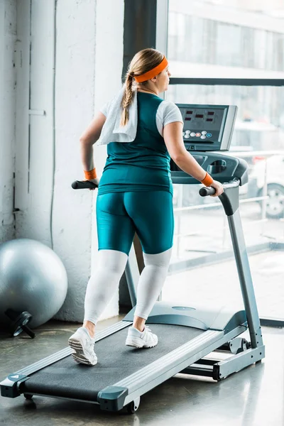 Overweight girl with towel on shoulders running on treadmill in gym — Stock Photo