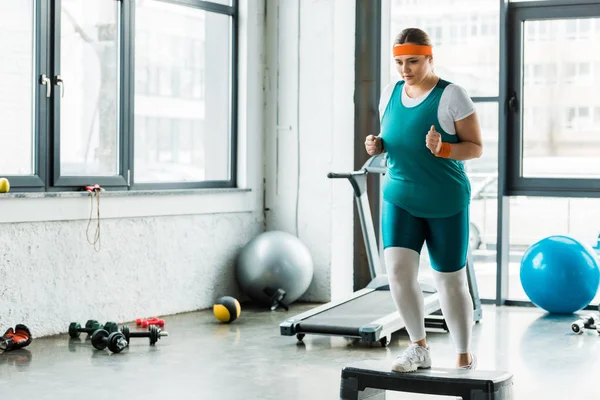 Плюс размер женщины упражнения на ступеньках платформы рядом со спортивным оборудованием — стоковое фото