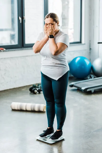 Overweight woman covering face with hands while standing on scales — Stock Photo