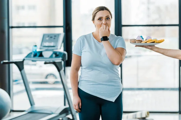 Vista recortada del hombre que sostiene la placa con pastelería sabrosa cerca de la mujer con sobrepeso que cubre la boca en el gimnasio - foto de stock