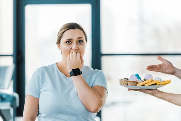 Vue recadrée de l'homme tenant plaque avec pâtisserie sucrée près de femme en surpoids couvrant la bouche dans la salle de gym — Photo de stock