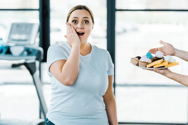 Vista ritagliata di uomo in possesso di piatto con gustosa pasticceria vicino sorpreso donna in sovrappeso in palestra — Foto stock