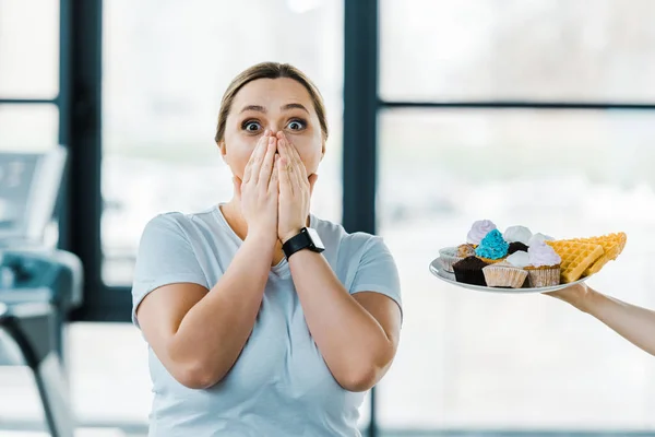 Vue recadrée de l'homme tenant la plaque avec la pâtisserie savoureuse près choqué femme en surpoids couvrant la bouche dans la salle de gym — Photo de stock