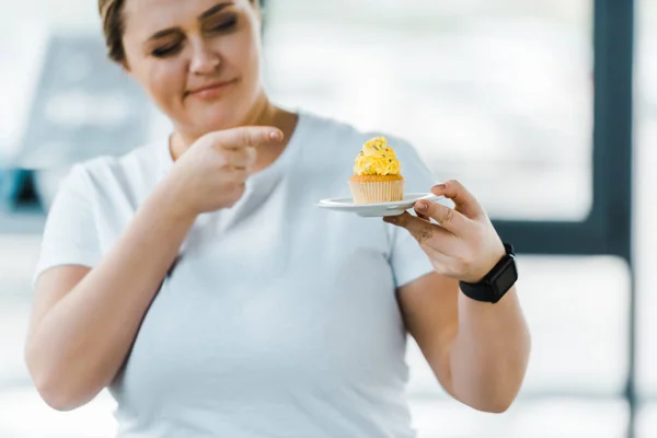 Cheerful overweight woman pointing with finger at tasty cupcake in gym — Stock Photo