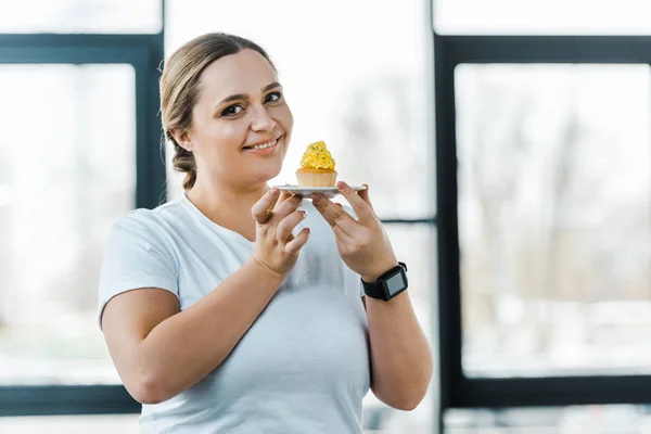 Heureuse femme en surpoids tenant délicieux cupcake dans la salle de gym — Photo de stock