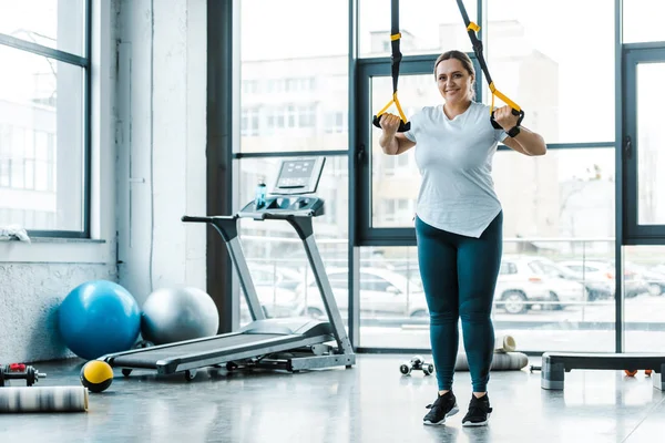 Fröhliche übergewichtige Frau trainiert Arme mit Spanngurten im Fitnessstudio — Stockfoto