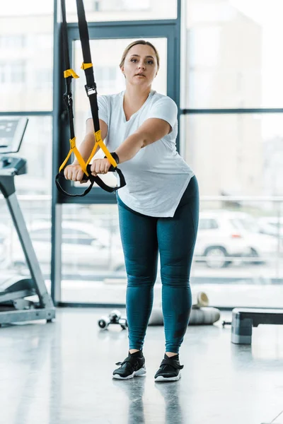 Overweight young woman training arms with suspension straps in gym — Stock Photo