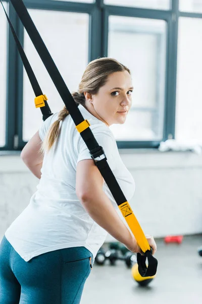 Attractive overweight woman training arms with suspension straps in gym — Stock Photo