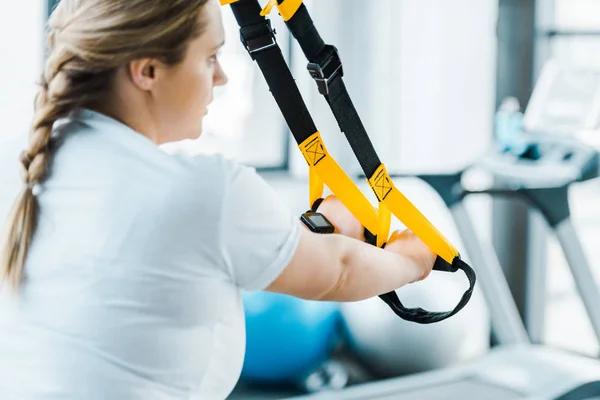Selective focus of overweight woman training arms with suspension straps in gym — Stock Photo