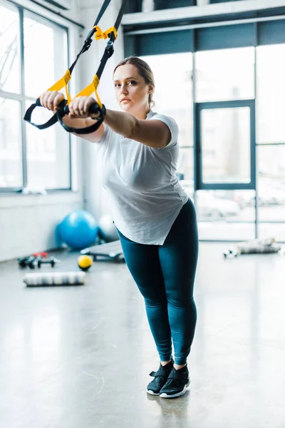 Concentrated overweight girl training arms with suspension straps in gym — Stock Photo