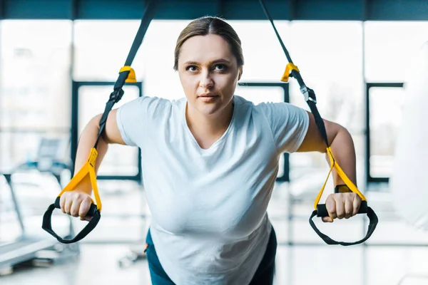 Konzentrierte Plus-Size-Mädchentrainingsarme mit Hängebändern im Fitnessstudio — Stockfoto