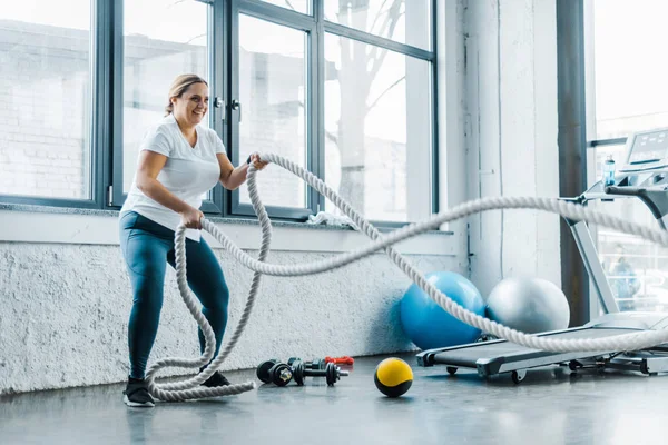 Alegre mujer con sobrepeso entrenamiento con cuerdas de batalla en el gimnasio — Stock Photo