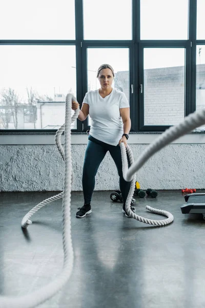 Concentrated overweight woman training with battle ropes in gym — Stock Photo