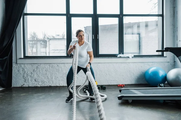 Entrenamiento de mujer de tamaño grande con cuerdas de batalla cerca de pelotas de fitness y cinta de correr en el gimnasio - foto de stock