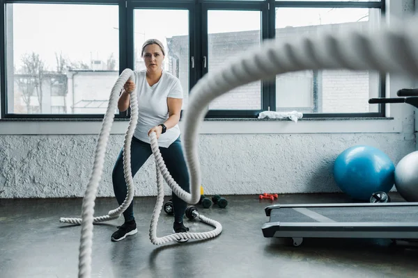Foco seletivo de treinamento focado plus size mulher com cordas de batalha no ginásio — Fotografia de Stock