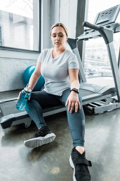 Fatigué plus taille femme reposant sur tapis roulant et tenant bouteille avec de l'eau — Photo de stock