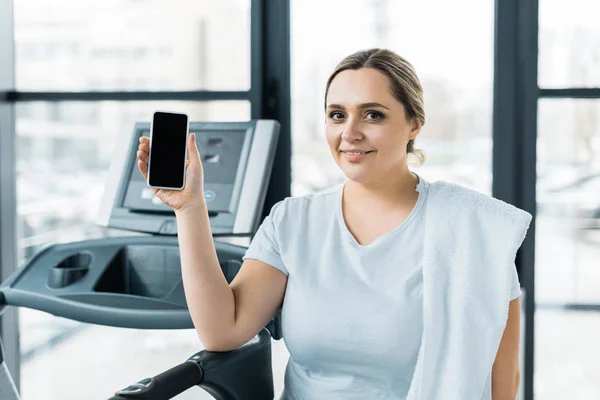 Alegre chica con sobrepeso sosteniendo teléfono inteligente con pantalla en blanco en el gimnasio - foto de stock